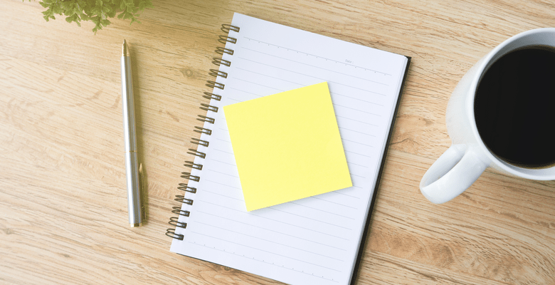 Auf einem Holztisch liegt ein Heft, ein Post-It, ein Kugelschreiber und eine Tasse Kaffee