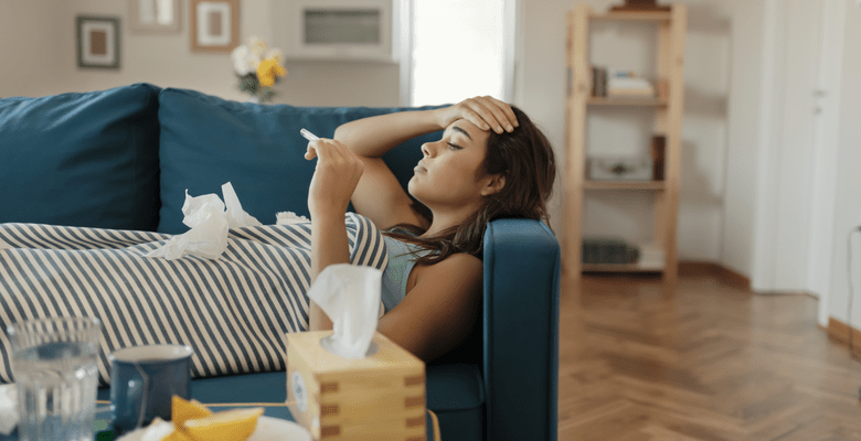 Eine Frau liegt auf der Couch mit einem Thermometer in der Hand, die andere Hand auf der Stirn