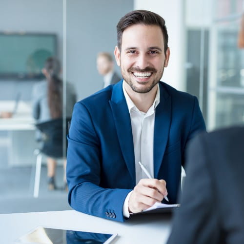 Mann im Business-Look, im Hindergrund ein moderner Besprechungsraum mit Menschen